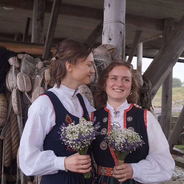 To kvinner i bunad smiler med blomsterbuketter