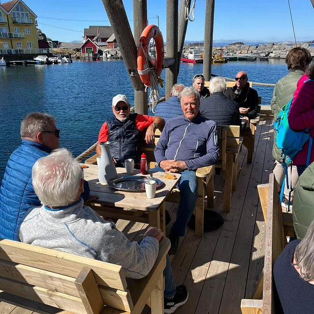 Folk sitter ute ved trebord ved sjøen og nyter solen